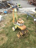 Rototiller shown outdoors on grass, visible rust on engine and metal parts, two wheels present, handlebar with rubber grips.