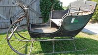 Side view of a vintage wood sleigh with a curved metal runner base showing wear and weathering on the wood paint, set outdoors on grass near wooden fencing.