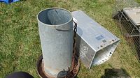 Cylindrical galvanized metal smoker with rusted chain and round base plate beside rectangular metal feeder outdoors on grass.