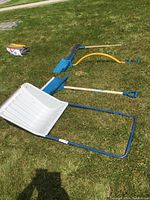 Photo showing all four items on grass: large white plastic scoop with blue frame, blue metal shovel with wooden handle, orange handled plastic shovel, and a blue plastic shovel with wooden handle.