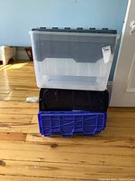 Stack of three plastic storage totes: clear with black lid, black tote with lid, and blue tote without lid.