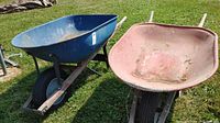 Two wheelbarrows on grass, one blue metal and one red metal with wooden handles and black tires.