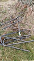 Two black metal log racks placed on grass near a tree, showing curved hooks and wear including rust.