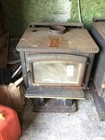 Front angled view of heavy steel woodstove showing glass door with metal trim and wooden handle, surface rust on top and front, vent pipe collar on top.