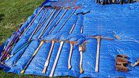 Assorted yard tools arranged on a blue tarp on grass including axes, pickaxe, rakes, hoes, pruners, and hedge trimmers.
