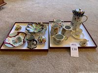 Two trays with various porcelain items including decorative bowls, plates, teapot, pitchers, cups, and a coffee pot. Items feature floral motifs, gold accents, and Victorian-style artwork.