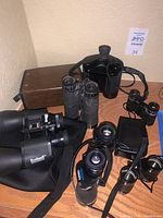 Overall view of multiple binoculars including a Bushnell large model with strap, smaller black textured binoculars, and carrying cases.
