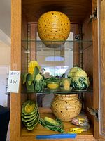 Front view of glass cabinet shelf displaying entire ceramic corn motif kitchenware lot including large corn basket, cookie jars, dishes, and napkin rings.
