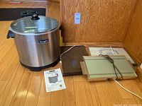 Charmglow stainless steel electric turkey fryer with glass lid and instruction manual, placed on hardwood floor next to three different vintage warming trays, one with green glass top and one with a wood or wood-like finish.