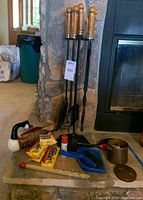 Four metal fireplace tools on a stand with wooden handles, goose-shaped holder with long matches, various fire starters and matchbooks on stone hearth.