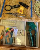 Drawer top view showing various utility items including magnifying glass, jar openers, flashlight, scissors, office supplies in drawer trays