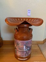 Full view of brown painted metal milk jug with attached tractor seat; shows the Budweiser saloon scene graphics and base text.