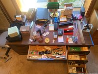 Overview of desktop with various office items arranged including desk lamp, stationery, pen holders, paper pads, and drawers partially open showing more supplies