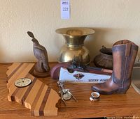Lot of Western decor items placed on a wooden surface, including carved wooden quail, wooden California block clock, brass spittoon, copper kettle, decanter shaped like a Kentucky flintlock rifle, decorative cowboy boot, and metal funnel.