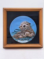 Framed artwork of two sea otters in a circular format with a wooden frame and black mat background.
