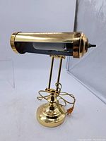 Front-right angled view showing the cylindrical brass shade, adjustable arm, round weighted base, power cord and overall polished brass finish.