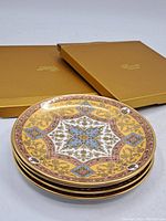 Stack of four ornate bone china porcelain side plates with gold, blue, and pink decorative pattern, placed next to two golden Fairmont Empress boxes.