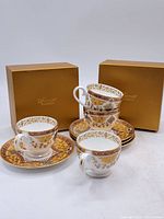 Photo shows four bone china teacups stacked with four matching saucers arranged with two gold color original boxes behind them.