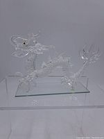 Clear crystal dragon figurine displayed on clear rectangular glass base, viewed from side showing full body and tail structure