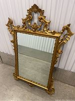 Full view of the gilded antique mirror standing against a wall, showing the rectangular reflective glass and the ornate gold frame with detailed scroll and leaf carvings.