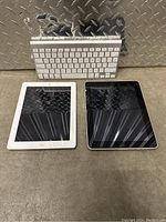 Two Apple iPads (white and black) alongside a wireless Apple keyboard on a gray floor with diamond plate backdrop.
