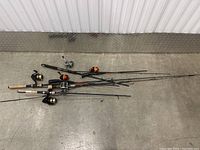 Full view of all five fishing rods laid out on a concrete floor, showing rods with reels and different handle designs.