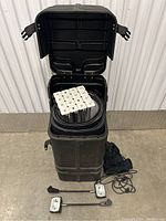 Carrying case standing upright with lid open, showing speaker system and two wired accessories placed in front.