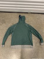 Single large green hoodie sweater laid flat on concrete floor showing front side with grey cuffs and hem accents and hood raised.