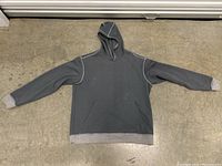 Grey hoodie sweatshirt laid flat on a concrete floor showing full front side with hood, kangaroo pocket, and light grey ribbed cuffs and waistband.