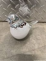 Front view of the Lasi Kostamo glass bird paperweight, showing clear glass head and tail, white body with brown and black speckles.