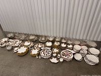 Wide shot showing stacks of plates, bowls, cups, saucers, and serving pieces arranged on floor against metal wall