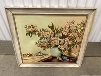 Front view of oil painting depicting a vase with flowers on a table, displayed in a white distressed wooden frame.
