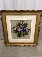 Framed needlework floral embroidery with detailed gold-tone decorative frame showing stitched purple flowers and green leaves.