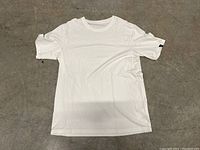 White Yeti crew neck T-shirt laid flat on a concrete floor, front side facing up. Shows the entire shirt clearly with short sleeves and crew neck style visible.