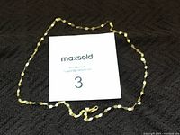 Photo of the 14k gold chain necklace laid out around a MaxSold card on a black textured surface, showing the full chain length and clasp area.