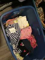 Blue plastic bin containing folded baby clothes including striped sleeper, pink polka dot garment, black pants with hearts, and other assorted tops and bottoms.