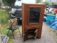 Full front view of the solid wood antique china cabinet with glass door, drawer and lower storage compartments, photographed outdoors near other items.