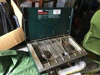 Front and top view of used Coleman propane camp stove showing two burners, control knobs, and metal grate with rust spots on surface.