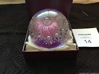 Front view of the clear glass crystal ball showing the pink flower design inside and bubbles, in an open display box with cream-colored cushion.