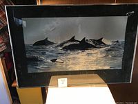 Framed large artwork showing dolphins swimming in water with reflective lighting, framed with black frame and glass, placed on a wooden surface.