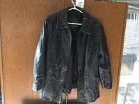 Front view of the black leather ladies medium coat hanging on a white hanger against a wooden door, showing the collar, sleeves, and overall condition of the coat.