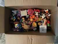Top view of open box filled with assorted miniature toys showing diverse shapes and colors including figurines and doll parts.