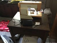 Singer sewing machine on wooden table with foot pedal and power cord visible.