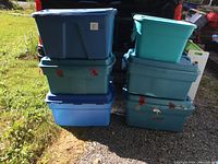 Six large plastic storage totes stacked in two columns. Various shades of blue and teal, all with lids, outdoors on grass and pavement.