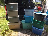 Stack of 11 large plastic storage totes with lids in various colors outdoors on grass and gravel surface.