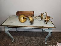 Light blue distressed wooden table with curved legs, two brass-colored multiple wall mount lamps on top, and wooden rectangular tray also on the table.