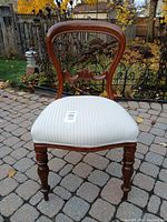 Front view of an antique balloon chair with wooden carved frame and white striped reupholstered seat cushion, set outdoors on cobblestone ground with autumn leaves in background.
