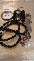 Photo showing four bracelets including black bead bracelets and one silver-tone patterned bangle, alongside two necklaces with pendants and a few pairs of silver-tone earrings.