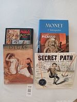 Five art books on a light surface. Titles visible include Pablo Picasso 1881-1973, Degas Impressions of a Great Master, Monet A Retrospective, Essential Picasso, and Secret Path graphic novel. All books fit coffee table size, largest approximately 10x14 inches. Condition shows minor wear on some covers.