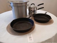 Two cast-iron frypans, large double boiler and two-piece stainless steel steamer pot arranged on a round woven placemat on table.